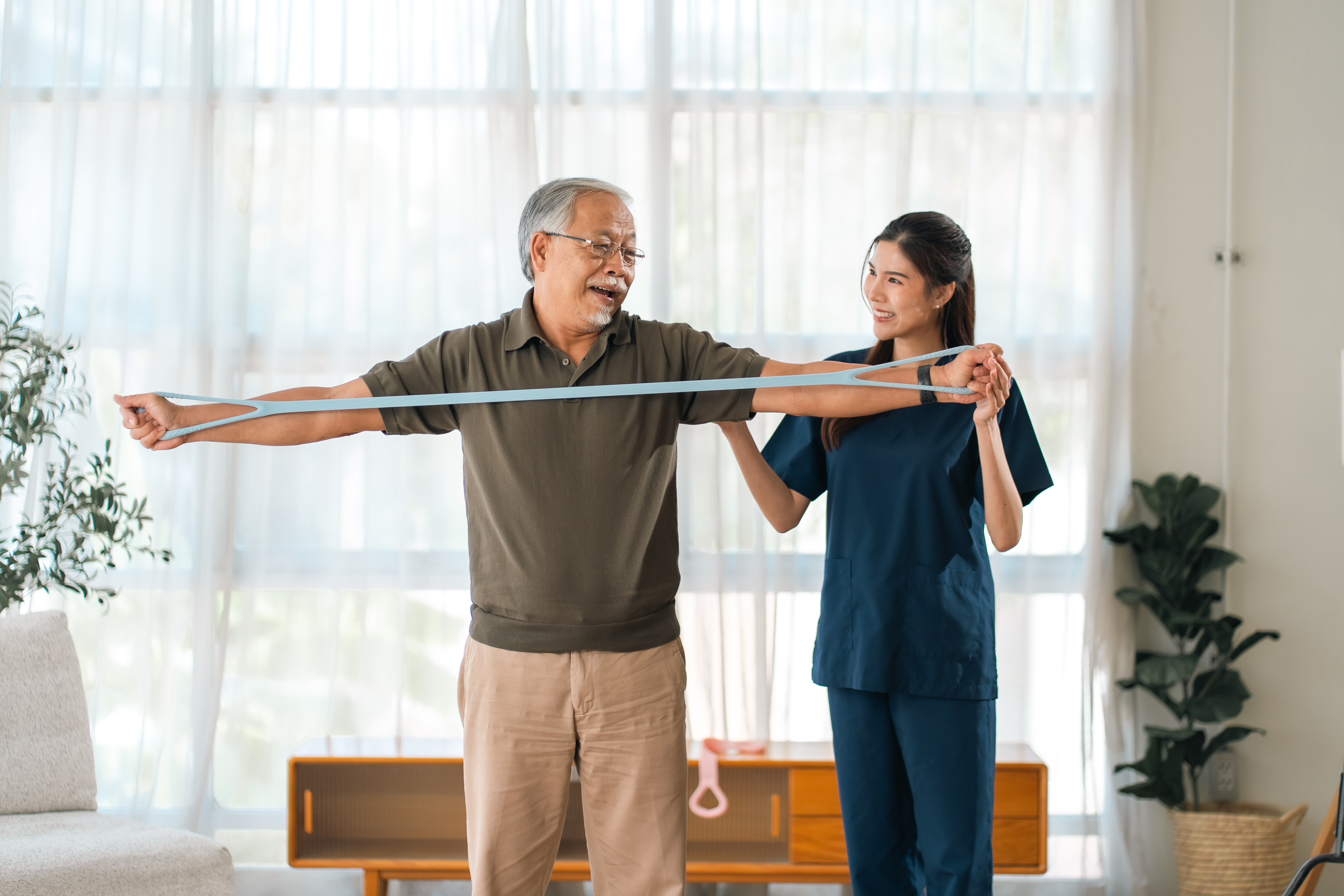 Caregiver with Asian senior patient, senior man and woman stretch therapy band in bright home wellness room